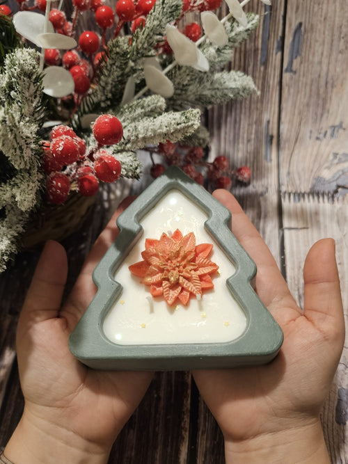Christmas Tree Candle held in hands, featuring a festive tree design with a red flower detail, surrounded by holiday greenery.